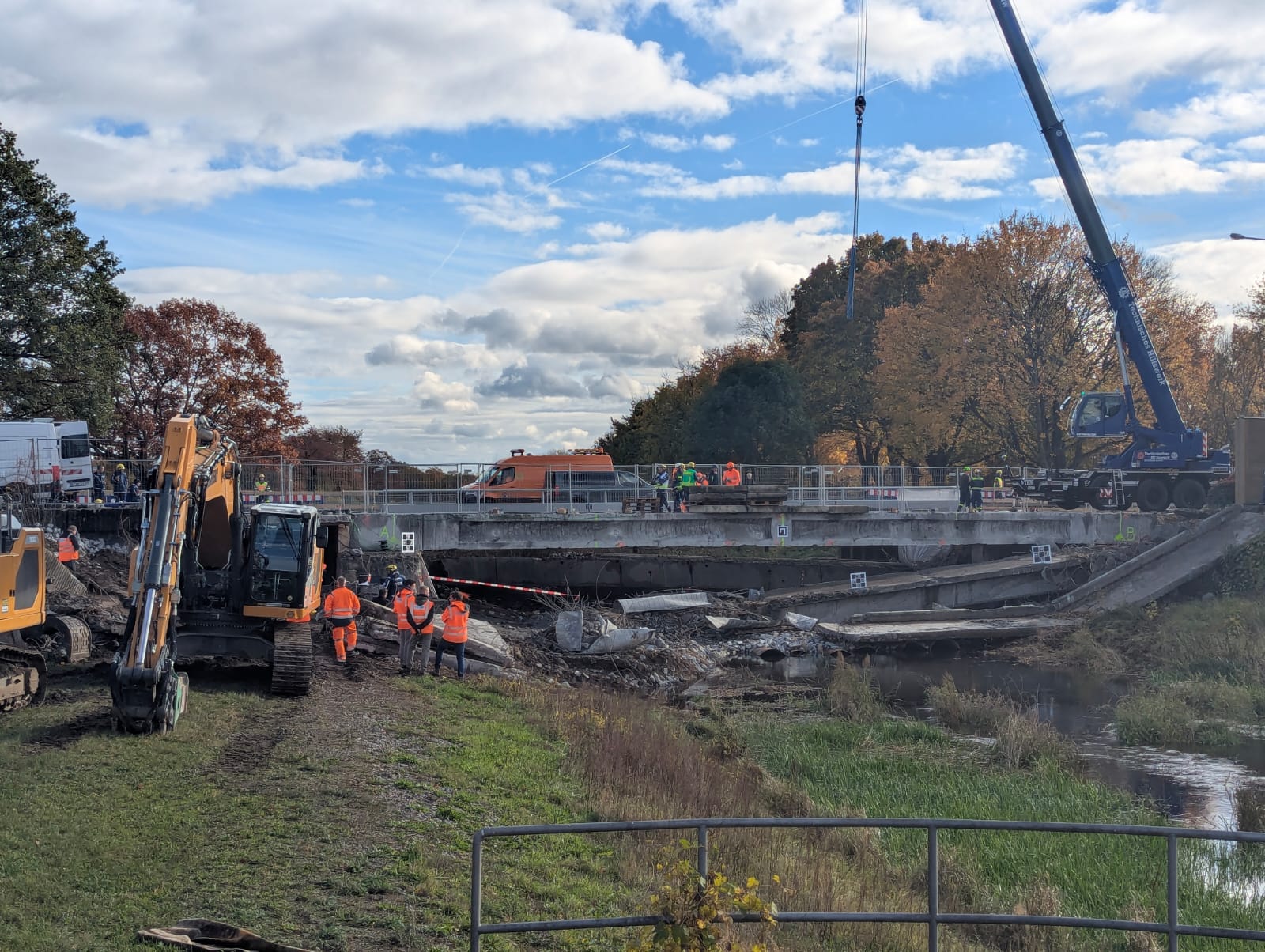 Bauingenieurin bzw. Bauingenieur (m/w/d) Bauüberwachung (QS Baustoffe/Abfall) Quaeransicht zur Brücke über den Fluss Schwarze Elster. Es sind Baumaschine, Bauarbeiter und Kontrolleure zu sehen. Ein blauer Kran steht restsseitig und hebt eine Betronplatte an. Rechts und links des Flusses befindet sich eine grüne Wieser. Der Himmel ist hellblau mit vielen weißen Wolken.