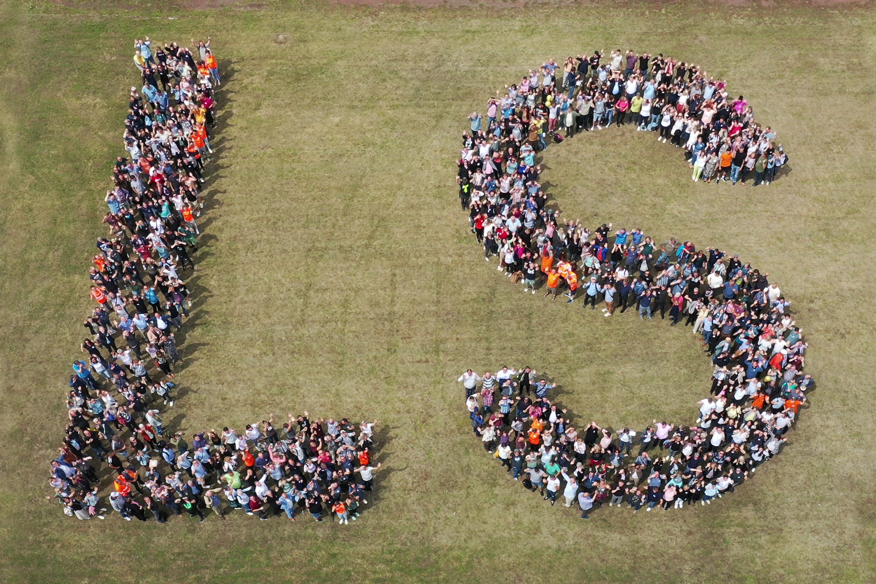 Über uns - Beschäftigte des Landesbetriebs Straßenwesen aufgestellt als die Buchstaben L und S. Der Untergrund ist eine grüne Wiesenfläche.