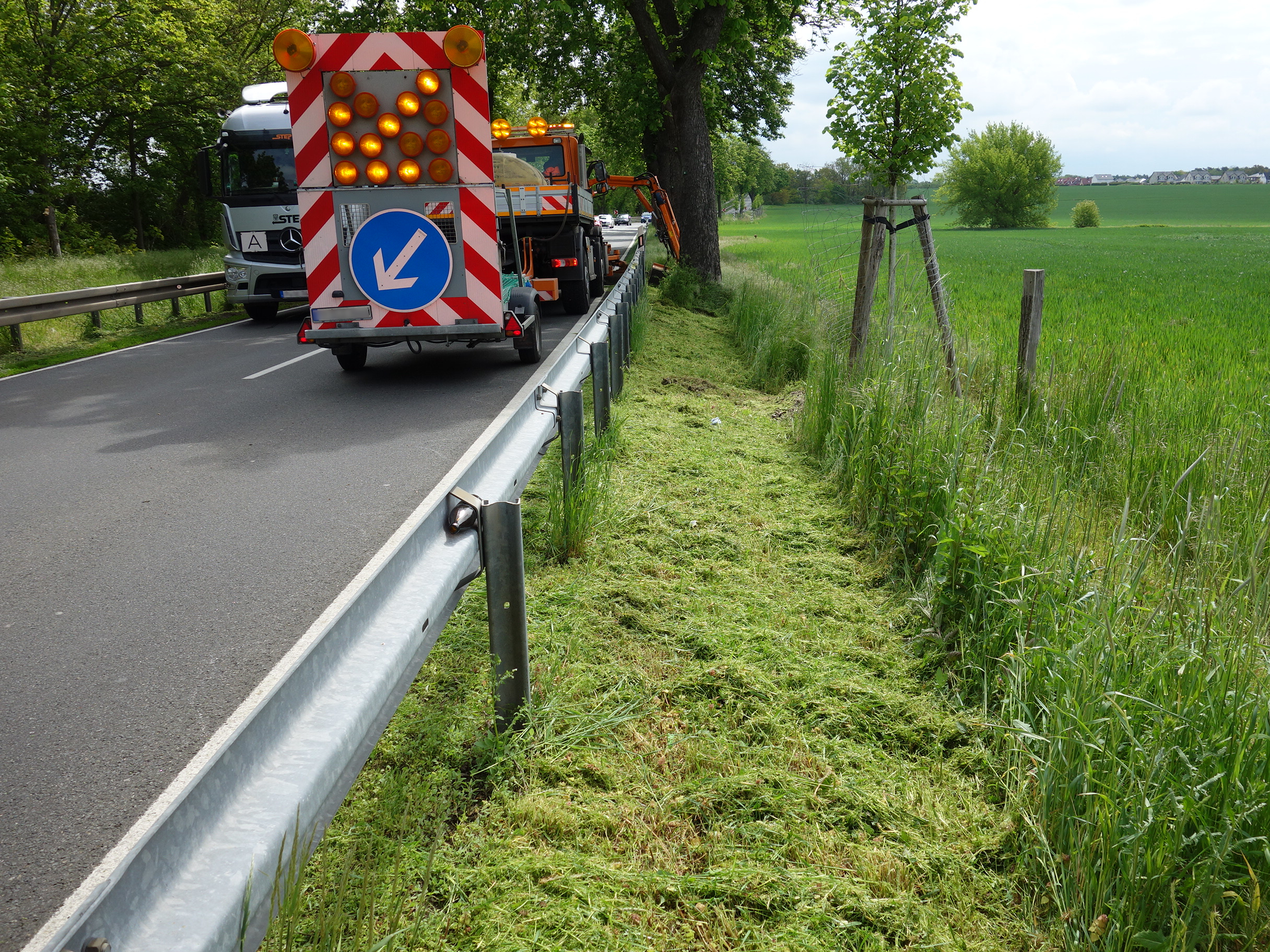Eine Allee mit Schutzplanken rechts und links, Ein Betriebsdienst-LKW der den Randstreifen mäht mit einem einem Schilderanhänger. Zu sehen oberhalb ein beleuchteter Richungspfeil nach links und darunden ein rundes blaues Verkehrsschild mit einem weißen Pfeil nach links ausgerichtet. Ein entgegenkommender LKW ist zur Häfte ebenfalls sichtbar.. Rechts neben der Straße ist ein grünen Feld.