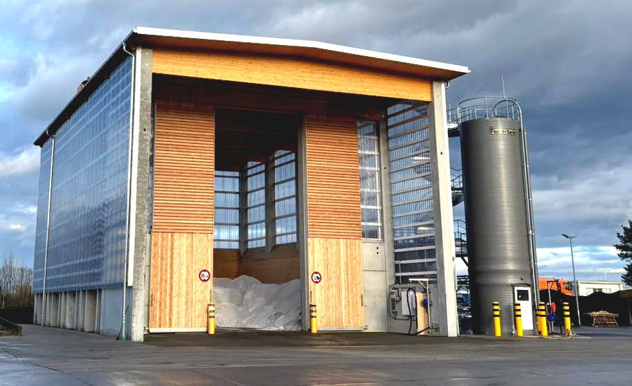 Auf einem grauen Betonuntersteht steht eine hohe Halle mit geöffneten Toren. Darin sieht man die Anfüllung von Streusalz. Rechts neben dem Bild steht ein Silo in dunkelgrau. Davor stehen 3 gelbe runde Poller mit 3 schwarzen Streifen obergalb der Poller.