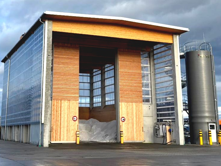 Auf einem grauen Betonuntersteht steht eine hohe Halle mit geöffneten Toren. Darin sieht man die Anfüllung von Streusalz. Rechts neben dem Bild steht ein Silo in dunkelgrau. Davor stehen 3 gelbe runde Poller mit 3 schwarzen Streifen obergalb der Poller.