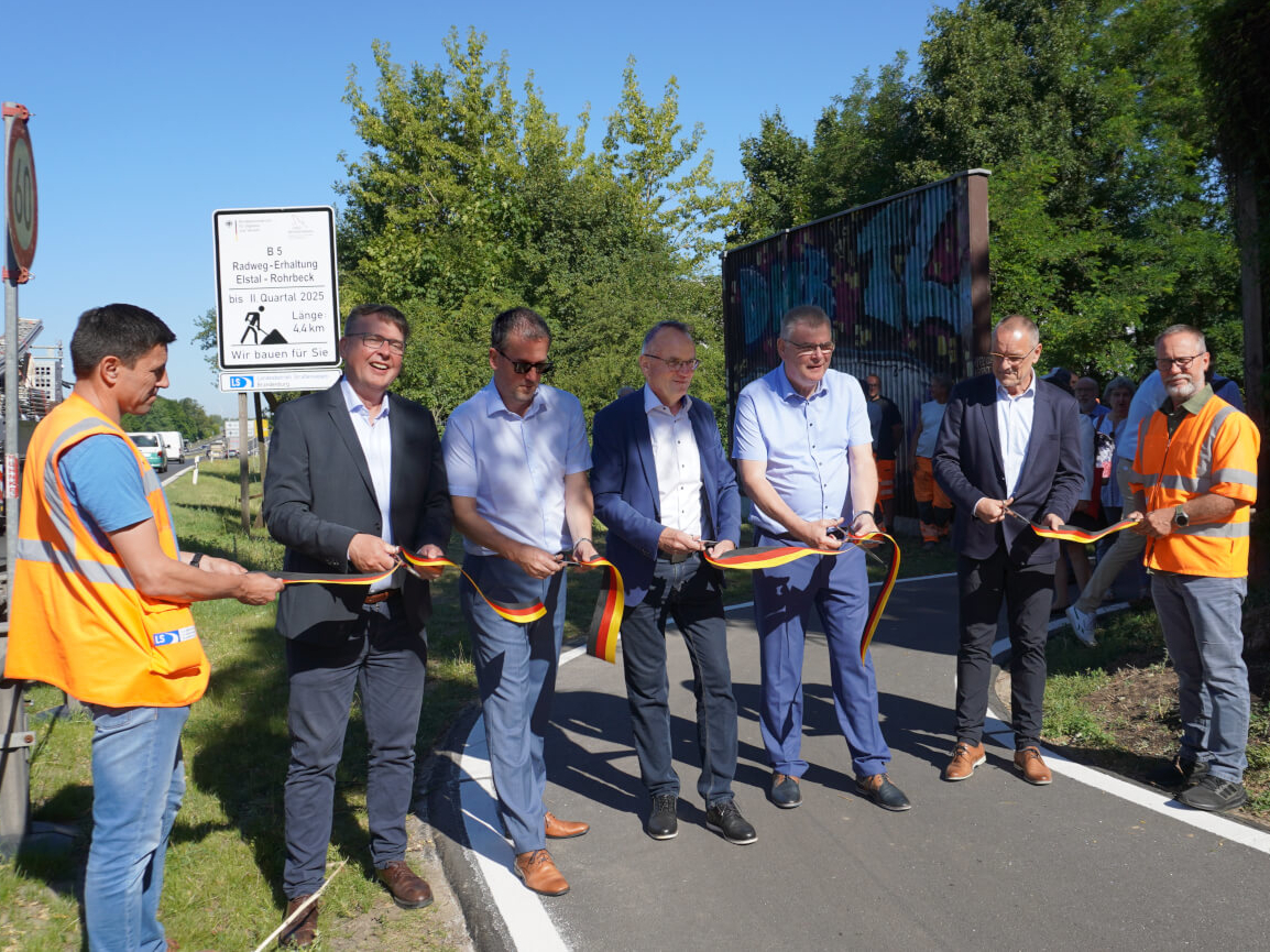 Das Band in den Farben schwarz rot gold wird zur Verkehrsfreigabe von einigen Gästen durchgeschnitten. In Hintergrund befindet sich das Baustellenschild.