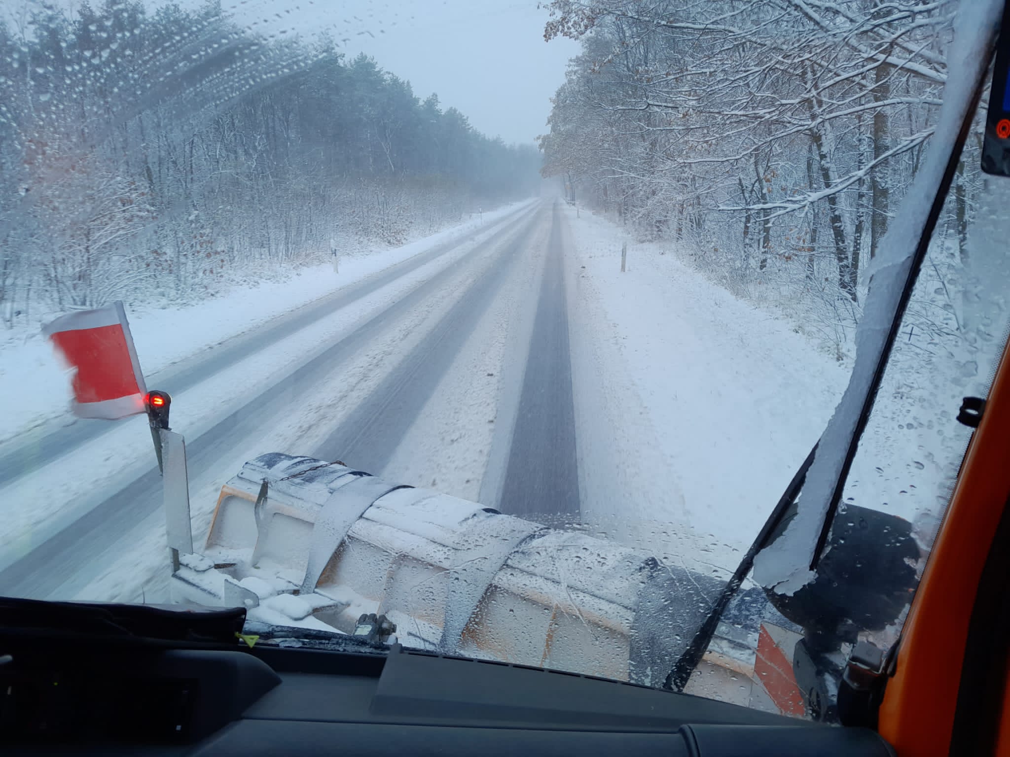 Blick aus dem Winterdienstfahrzeug vornau auf die Straße, die gerade beräumt wird. Rechts und links der Straße stehen schneebedeckte Bäume.