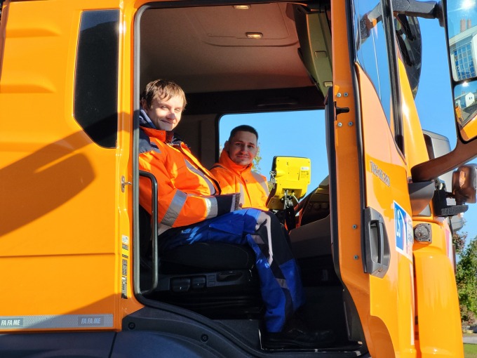 Blick von unten nach oben in eine orangsfarbenes Betriebsdienst-LKW. Die Beifahrertür ist geöffnet. Zu sehen sind Maik Wolff und Johannes Briesener, die in die Kamera lächeln.