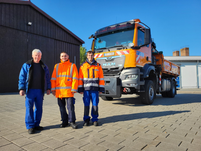 Das Foto zeigt einen Teil des Innenhofs der Straßenmeisterei Fürstenwalde. Der Untergrund besteht aus grauen Pflandersteinen. Im Hindergrund ist eine schwarue Halle zu sehen sowie ein Betriebsdienst-LKW der Firma MAN in orange. An der Fahrerseite ist das Loge des LS abgebildet. Im Vordergrund stehen von links nach rechts Thoralf Kaps aus der Straßenmeisterei, Mail Wolff und Johannes Briesner.