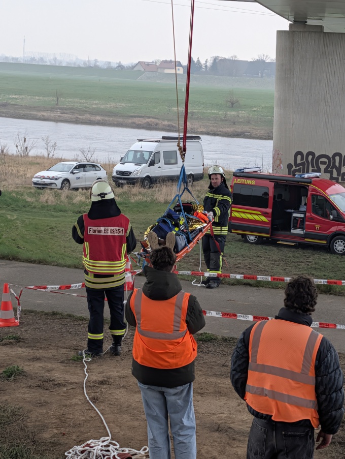 Rettungsübung Bergung eines Verletzten. Im Hintergrund sind 2 weiße Sienstfahrzeuge zu sehen, dahinter die Elbe, Wiese und ein paar Häuser. Leichter Nebel liegt über der Wiese.