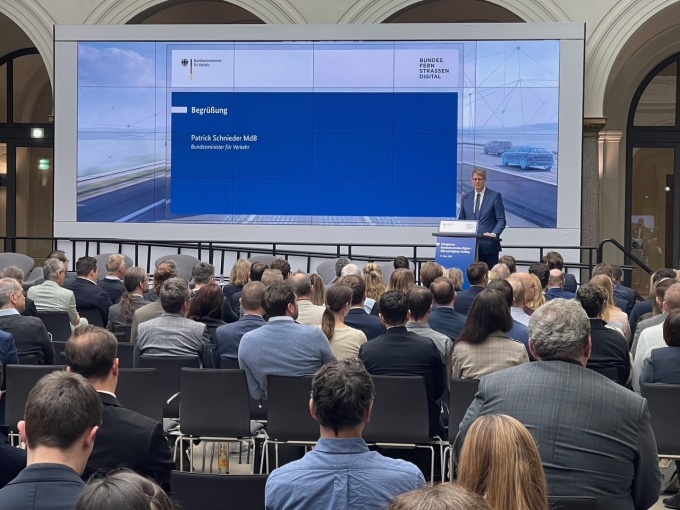 Im Vordergrund siht man sitzende Menschen von hinten, dahinter steht Bundesverkehrsminister Schnieder hinter einem blauen Rednerpult von einer blauenen Rückwand.