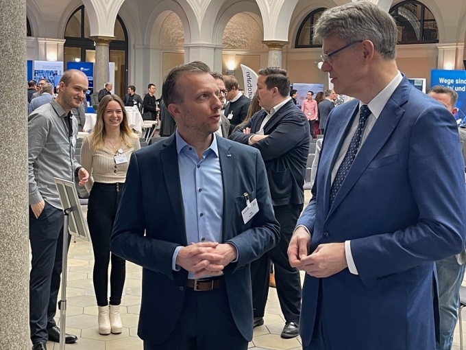 Mike Koehler Vorsitzender des LS spricht mit Bundesverkehrsmister Schnieder. Im Hintergrund sihr dan einige der Teilnehmer innerhalb des Veranstaltungsortes. von einer blauenen Rückwand.
