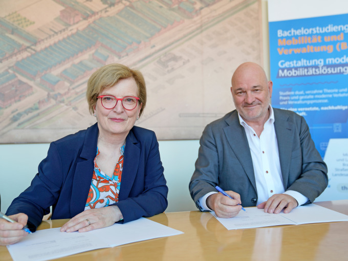 Präsidentin Ulrike Tippe und Minister Robert Crumbachsitzen an einem Tisch und schauen lächenld in die Kamera. Sie halten einen Stift in ihrer rechten Hand.Vor lihnen liegt jeweils der Kooperationsvertrag in Papierform.