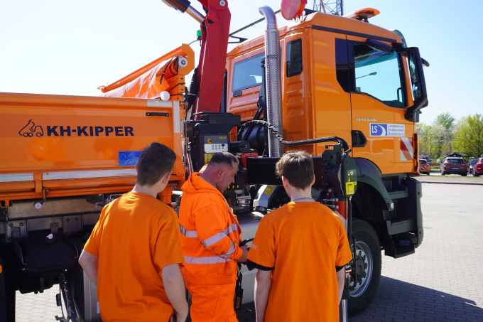 Im Hintergrund befindet sich ein orange farbener LKW ein Kipper von der Beifahrerseite. Auf der Beifahrertür ist das LS Logo zu sehen. Vor dem Fahrzeug steht ein Beschäftigter der Straßenmeisterei Rehfelde mit 2 Schülern. Alle sind orange gekleidet.